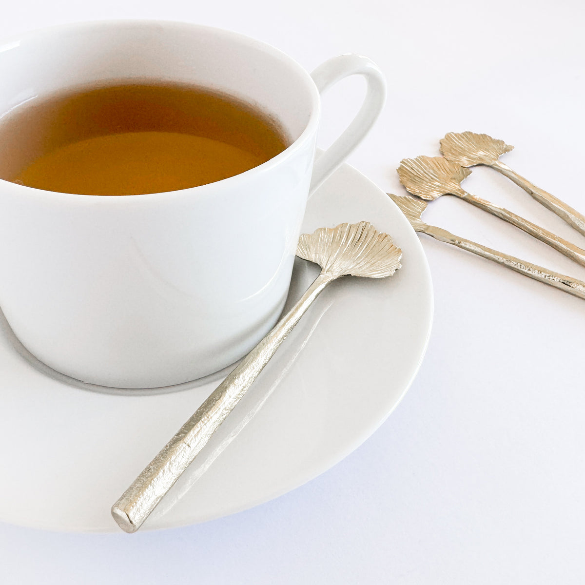M. By Design - Curated tabletop essentials. White cup of tea with a decorative spoon on a white plate, set against a white background.